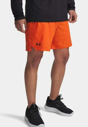 Pantalones cortos deportivos de color naranja brillante con bolsillos laterales y un logo negro de Under Armour. Combinados con zapatillas deportivas negras con una suela gris.