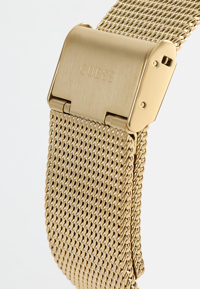 Bracelet de montre en mailles dorées avec un fermoir lisse et poli portant le mot "GUESS" gravé. Le motif en mailles est serré et uniformément espacé.