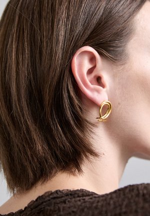 Goldene Creolen mit einem glatten, verdrehten Design; vor kurzen braunen Haaren präsentiert, die ihren Glanz und ihre Form betonen.