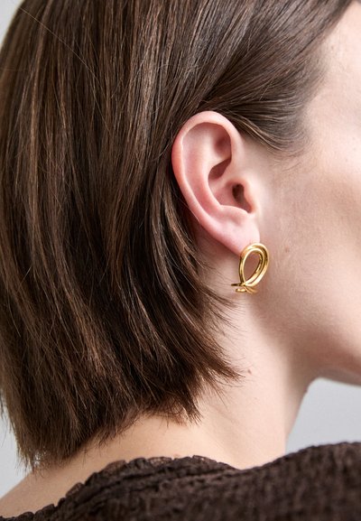 Aro de oro con un diseño liso y retorcido; exhibido contra cabello corto castaño, destacando su brillo y forma.