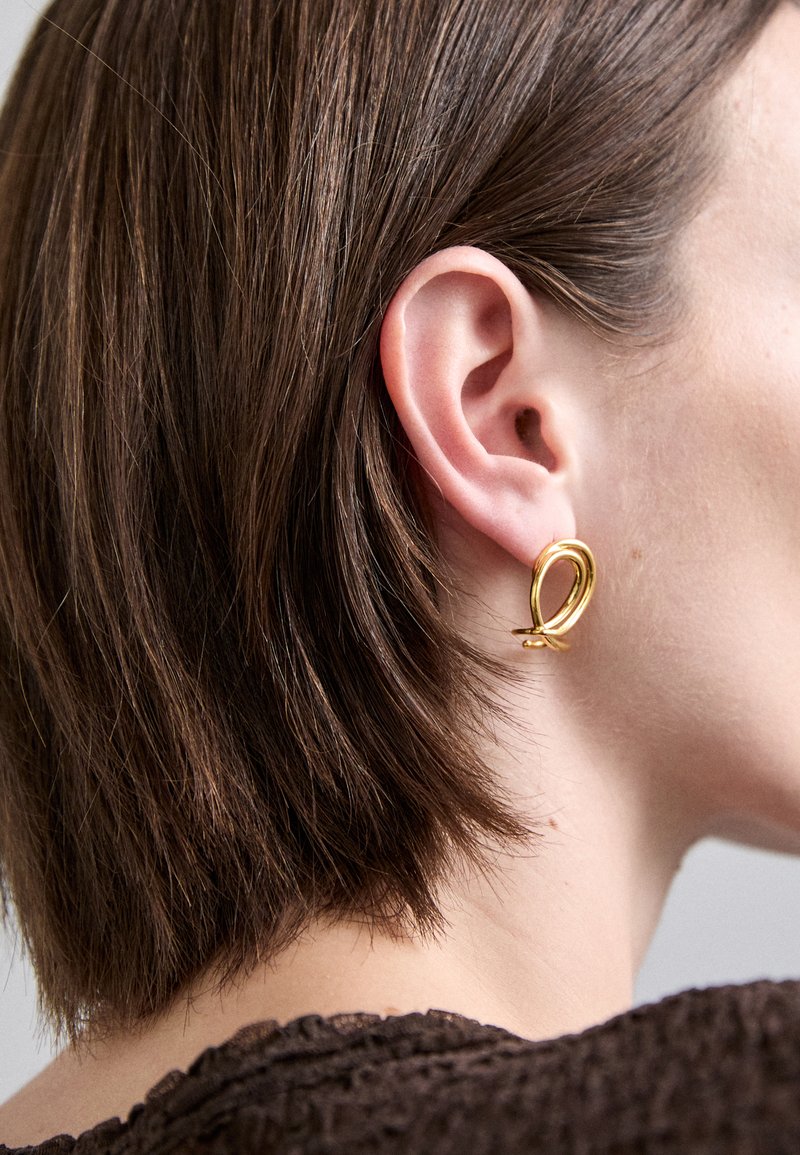 Złota kolczyk w kształcie obręczy o gładkim, skręconym wzorze; wyeksponowany na tle krótkich brązowych włosów, podkreślający jego blask i kształt.