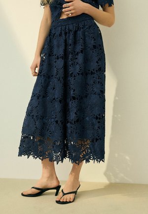 Frau trägt marineblauen floralen Spitzen-Midi-Rock und passendes Oberteil, kombiniert mit schwarzen Sandalen mit Absatz und Zehensteg, Hand ruht in der Nähe der Taille.
