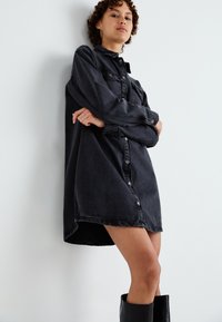 Zwarte denim blousejurk met lange mouwen, een licht gebogen zoom en accenten in zilver. Model staat tegen een lichte wand.