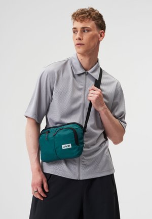 Junger Mann trägt ein hellgraues kurzärmeliges Reißverschlusshemd, schwarze Hose, hält eine grüne Umhängetasche mit schwarzem Riemen über der Schulter.
