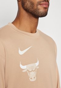 Bež majica kratkih rukava s okruglim izrezom. Ima bijeli Nike swoosh i grafiku bika na prednjem dijelu. Mekana tekstura, ležeran dizajn.