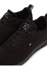Paire de baskets en maille noire Tommy Hilfiger avec lacets noirs, étiquettes de languette de marque et petit logo rouge, blanc et bleu sur les côtés et sur les languettes de talon.