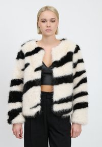 Faux fur-jakke med sort og hvide striber, der har en blød tekstur, åben front og brede ærmer. Båret over en sort top og bukser.