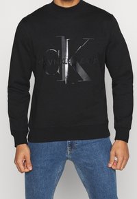 Uomo che indossa un felpa nera Calvin Klein con logo in rilievo e jeans blu, in piedi contro uno sfondo leggero e uniforme.