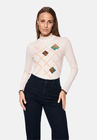 Langärmliges cremefarbenes Oberteil mit Rüschenausschnitt, verziert mit lebhaften orangefarbenen und gemusterten Diamantdesigns, kombiniert mit dunkelblauen Denim-Hosen.