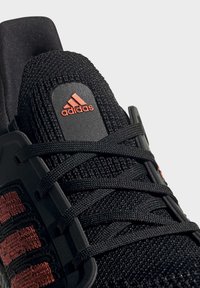 Chaussure de sport noire avec une tige texturée, des accents orange, un système de laçage, et une semelle extérieure noire proéminente avec un logo sur la languette.