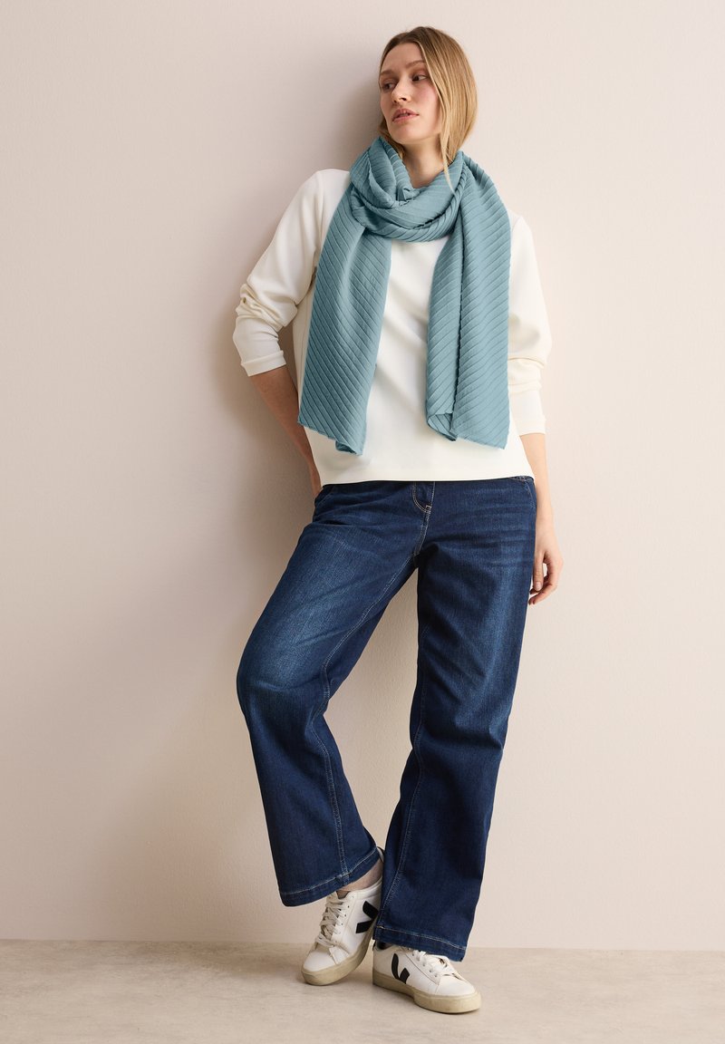 Écharpe texturée bleu clair, haut à manches longues blanc, jeans évasés bleu foncé, et baskets blanches avec des accents noirs. Debout contre un mur neutre.