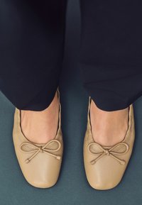 Beige nahka ballerina-kengät pyöreillä varpailla, edessä koristeena sulka ja näkyvät ompeluyksityiskohdat reunoilla.
