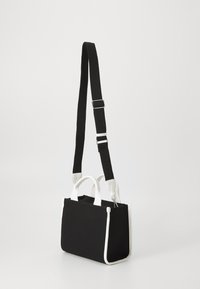 Sac en toile noir avec des accents synthétiques blancs, forme rectangulaire, deux poignées, et une bandoulière amovible pour plus de polyvalence.