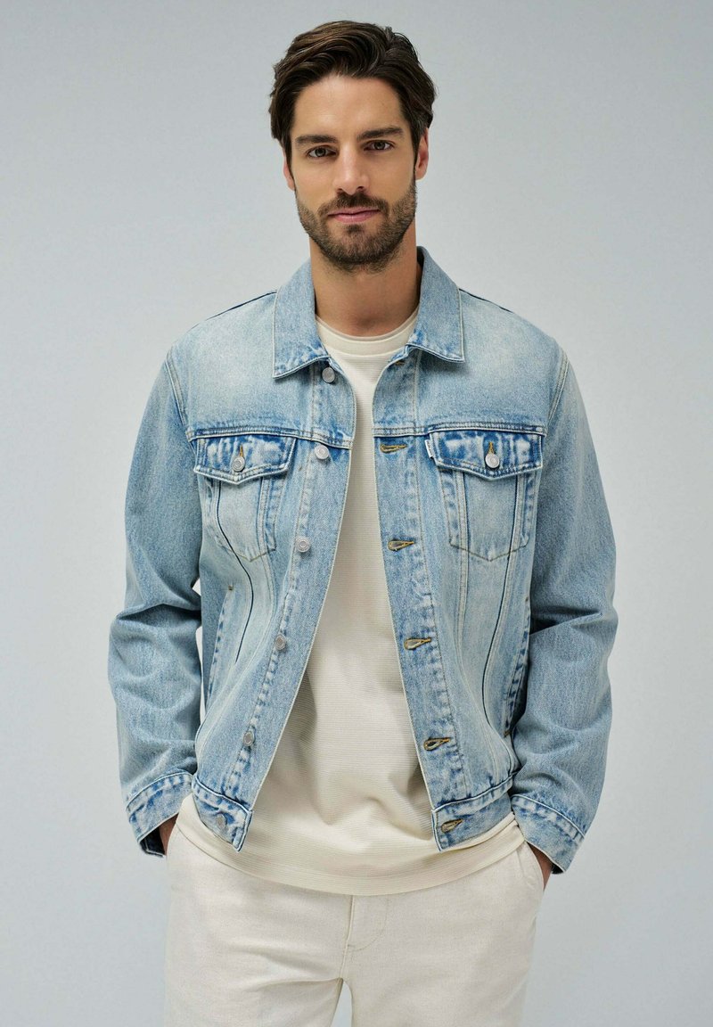 Hellblaues Denim-Jacket mit klassischem Design, ausgestattet mit zwei Brusttaschen, Knopfschluss und einer leicht ausgebleichten Textur. Getragen über einem beigen Oberteil.