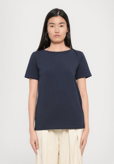 T-shirt bleu marine à manches courtes avec un col rond, fabriqué en tissu doux. Design simple, coupe droite, porté avec un pantalon plissé de couleur claire.