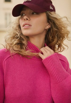 Eheline roosa kootud sviiter kõrge kraega ja soonikkoes mansettidega. Mudel kannab tumedat burgundi värvi nokamütsi, mis rõhutab pehmeid, lokkis juukseid.