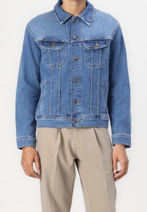 Uomo che indossa una giacca di jeans blu con chiusure a bottoni e tasche sul petto con patta, abbinata a pantaloni beige plissettati.