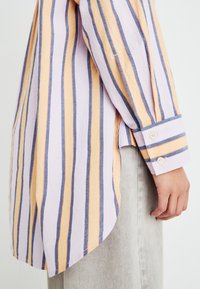 Chemise légère à rayures dans des tons de violet, orange et bleu. Dotée d'un ourlet arrondi et de poignets à boutons. Tissu doux et texturé.