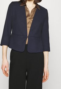 Blazer bleu marine à coupe ajustée, épaules arrondies, manches trois-quarts, et fermeture avant unique, associé à une chemise en satin marron.