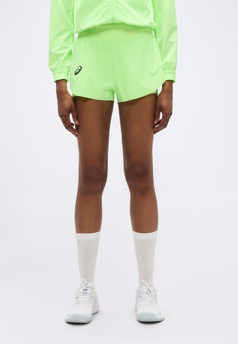 Άτομο που φοράει αθλητικά σορτς και τοπ σε νέον πράσινο χρώμα, λευκές κάλτσες πλήρους μήκους και λευκά παπούτσια τρεξίματος, στέκεται σε κανονικό λευκό φόντο.