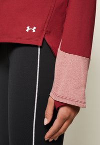 Gros plan d’une main portant un haut de sport à manches longues rouge et rose avec un trou pour le pouce, appuyée contre des leggings noirs avec une bande verticale blanche.