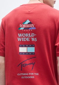 Camiseta de algodão vermelha com design gráfico apresentando o logótipo "tommy jeans", silhueta de montanha e texto "WORLD-WIDE '85" nas costas.