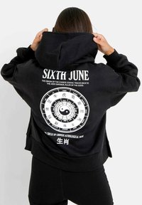 Felpa nera con cappuccio caratterizzata da una grafica circolare bianca con simboli zodiacali cinesi e testo. Il design include un simbolo yin-yang al centro.