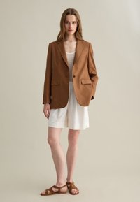 Blazer in lino marrone con rever a punta, chiusura a un bottone e tasche frontali, abbinato a una canotta crema e pantaloni corti bianchi.