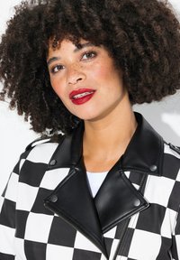 Veste en simili cuir à carreaux noir et blanc avec un col matelassé noir, des accessoires en acier inoxydable et une fermeture éclair. Détail de surface texturé présent.