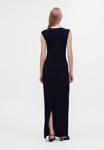 Femme aux longs cheveux blonds portant une robe maxi noire sans manches avec une fente dans le dos et des chaussures noires à talons bas, debout sur un fond blanc.