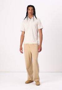 Polo beige a maniche corte, abbinato a pantaloni larghi di un beige chiaro. Il tessuto appare liscio e leggero, adatto per un abbigliamento casual.
