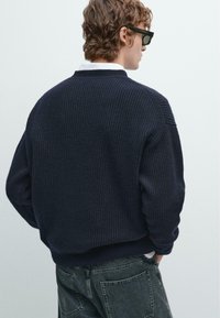 Maglione a coste navy con una vestibilità rilassata, scollo rotondo e superficie texturizzata, abbinato a jeans scuri. Dettagli sottili sulle cuciture delle spalle.