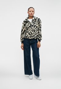 Fleece-Pullover in Schwarz und Creme mit abstrakten Mustern, Reißverschlusskragen und Fronttasche. Kombiniert mit weiten, dunklen Jeans und weißen Sneakers.