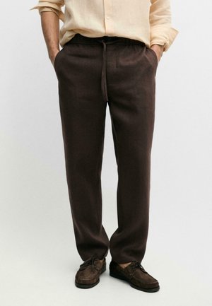 Uomo che indossa pantaloni marrone scuro con coulisse, camicia beige a maniche lunghe con maniche arrotolate e scarpe casual marrone scuro.