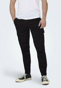 Pantalons cargos noirs avec des poignets élastiques, présentant plusieurs poches latérales. Portés avec un t-shirt blanc et des baskets noires. Texture de tissu lisse.