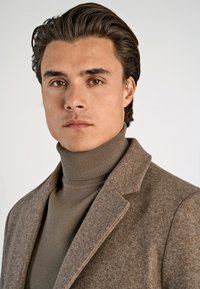 Manteau en laine marron avec des revers crantés, associé à un col roulé kaki. Les textures lisses et la coupe ajustée mettent en avant un look raffiné.