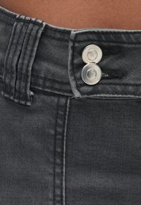 Mustast teksariidest kangas, millel on vöökohal lähedalt vaadatuna kaks hõbedast nööpi ja nähtavad õmbluse detailid. Pehme tekstuur ja struktureeritud disain.