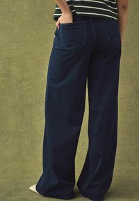 Weite, dunkelblaue Denim-Hosen mit klassischem Fünf-Taschen-Design. Glatte Textur mit sichtbaren Nähten und subtilen Details an den Gesäßtaschen.