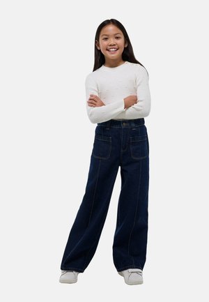 Jeans en denim foncé à jambe large avec poches avant et coutures apparentes, associés à un haut à manches longues côtelé blanc. Modèle souriant avec les bras croisés.