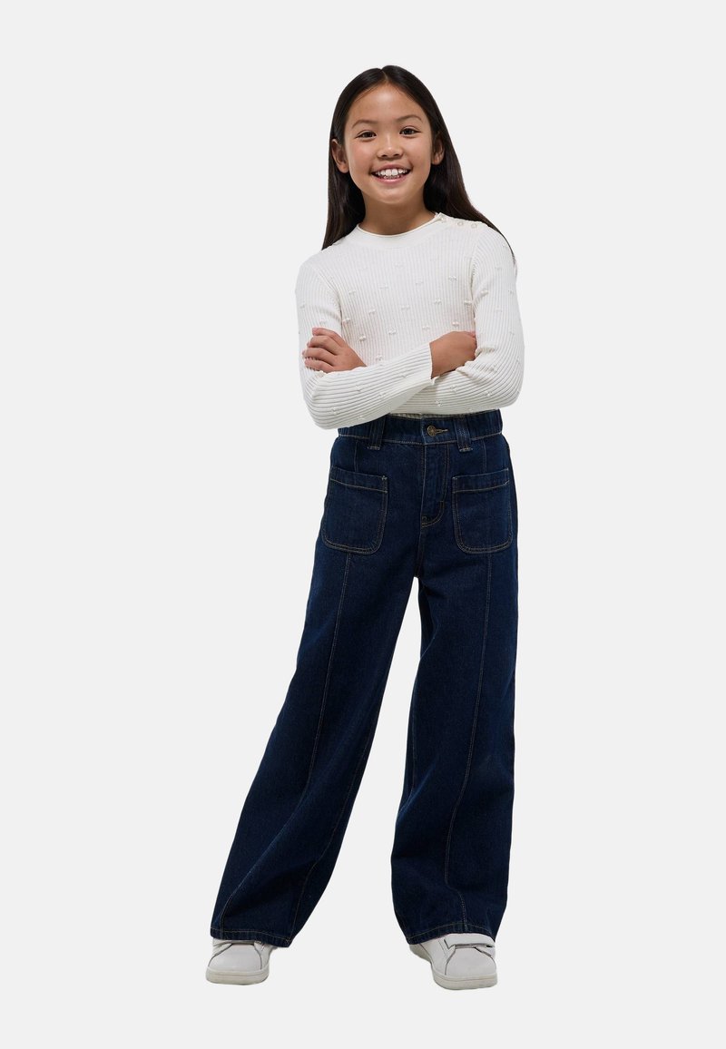 Jeans en denim foncé à jambe large avec poches avant et coutures apparentes, associés à un haut à manches longues côtelé blanc. Modèle souriant avec les bras croisés.
