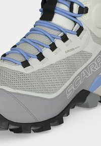 Gros plan d'une chaussure de randonnée grise et blanche avec des lacets bleus, une tige en mesh respirant, une semelle noire robuste et le logo « SCARPA » sur le côté.