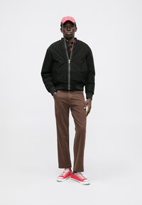 Černá bombardérka, košile s červeným a černým kostkovaným vzorem, hnědé kalhoty, červené tenisky, růžová čepice. Neformální streetwear styl s moderním střihem.