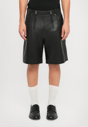 Mann in schwarzen Lederknielangen Shorts mit zwei vertikalen Vorderreißverschlüssen, weißen Sportsocken und schwarzen Lederhalbschuhen.