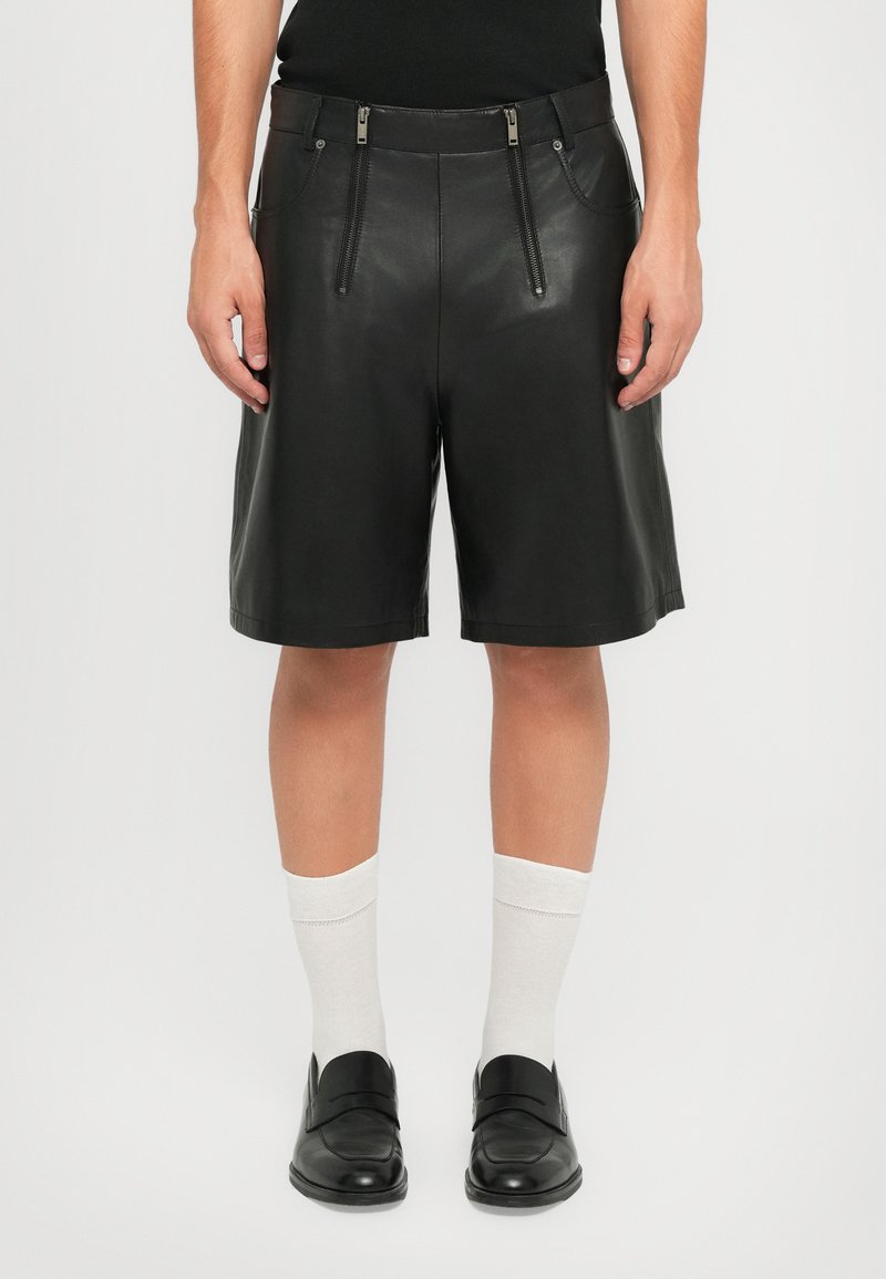 Man wearing black leather knee-length shorts with two vertical front zippers, white crew socks, and black leather dress shoes.