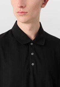 Černá košile s límečkem, jemnou pruhovanou texturou a pěti šedými knoflíky podél légu. Hladký povrch látky.