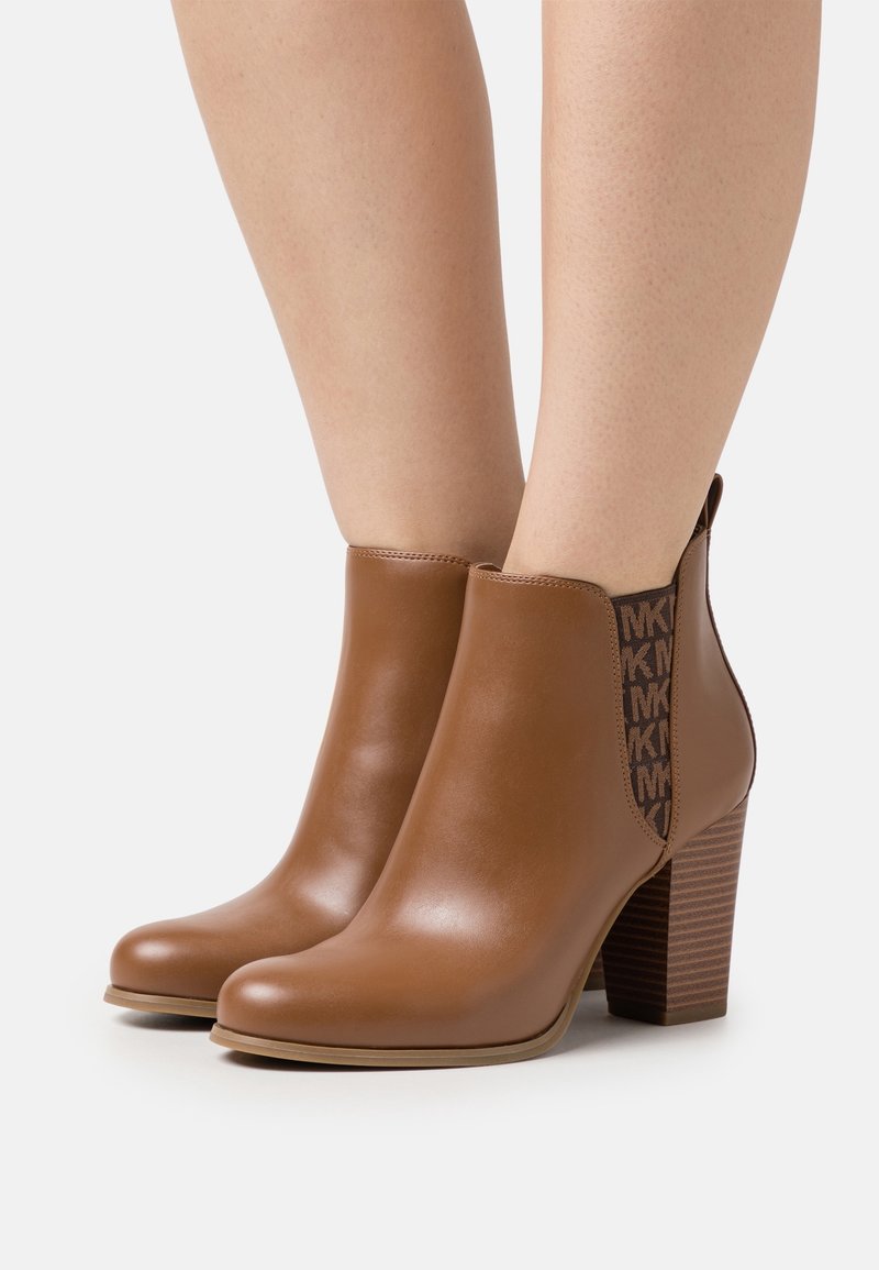 Bottines en cuir marron avec talons carrés empilés et panneaux latéraux élastiques affichant le logo "MK" en répétition, portées sur jambes nues.