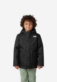 Černá nepromokavá bunda s kapucí, se zapínáním na přední straně, dvěma předními kapsami a bílým logem na levé hrudi, nošená přes světle zelené kalhoty.