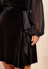 Schwarzes Wickelkleid mit samtiger Textur, langen durchsichtigen Ärmeln und einem Rüschensaum. Der Stoff reflektiert das Licht subtil.