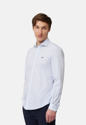 Giovane uomo con capelli castano chiaro che indossa una camicia a righe azzurra e bianca con bottoni e pantaloni scuri, in piedi davanti a uno sfondo semplice.