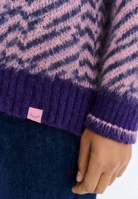 Zbliżenie na dłoń osoby obok mankietu swetra dzianinowego w fioletowo-różowy zygzakowaty wzór oraz ciemnoniebieskich spodni.